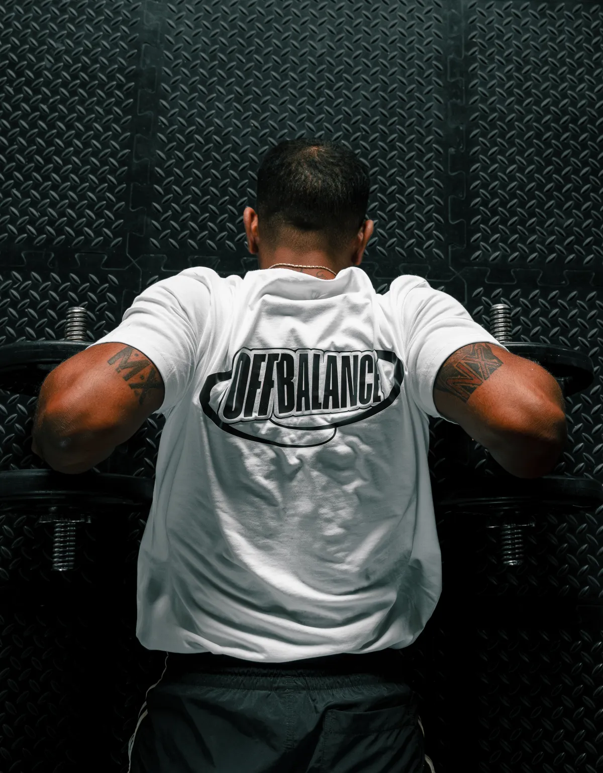 Off Balance coach mid-rep with dumbbells, back print facing the camera against a dark training wall
